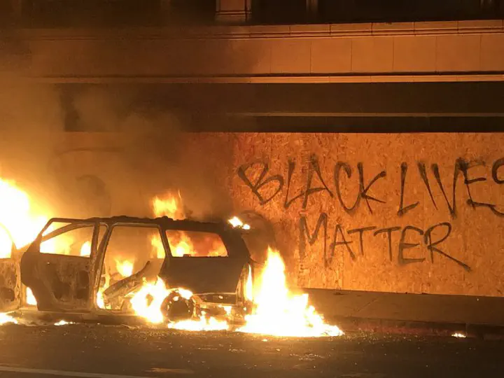 Burning vehicle with Black Lives Matter graffiti on wall behind